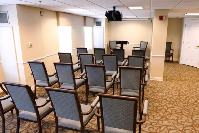 A small meeting or presentation room with rows of gray cushioned chairs facing a piano and a wall-mounted TV screen. The room has beige walls, patterned carpet, and windows letting in natural light.