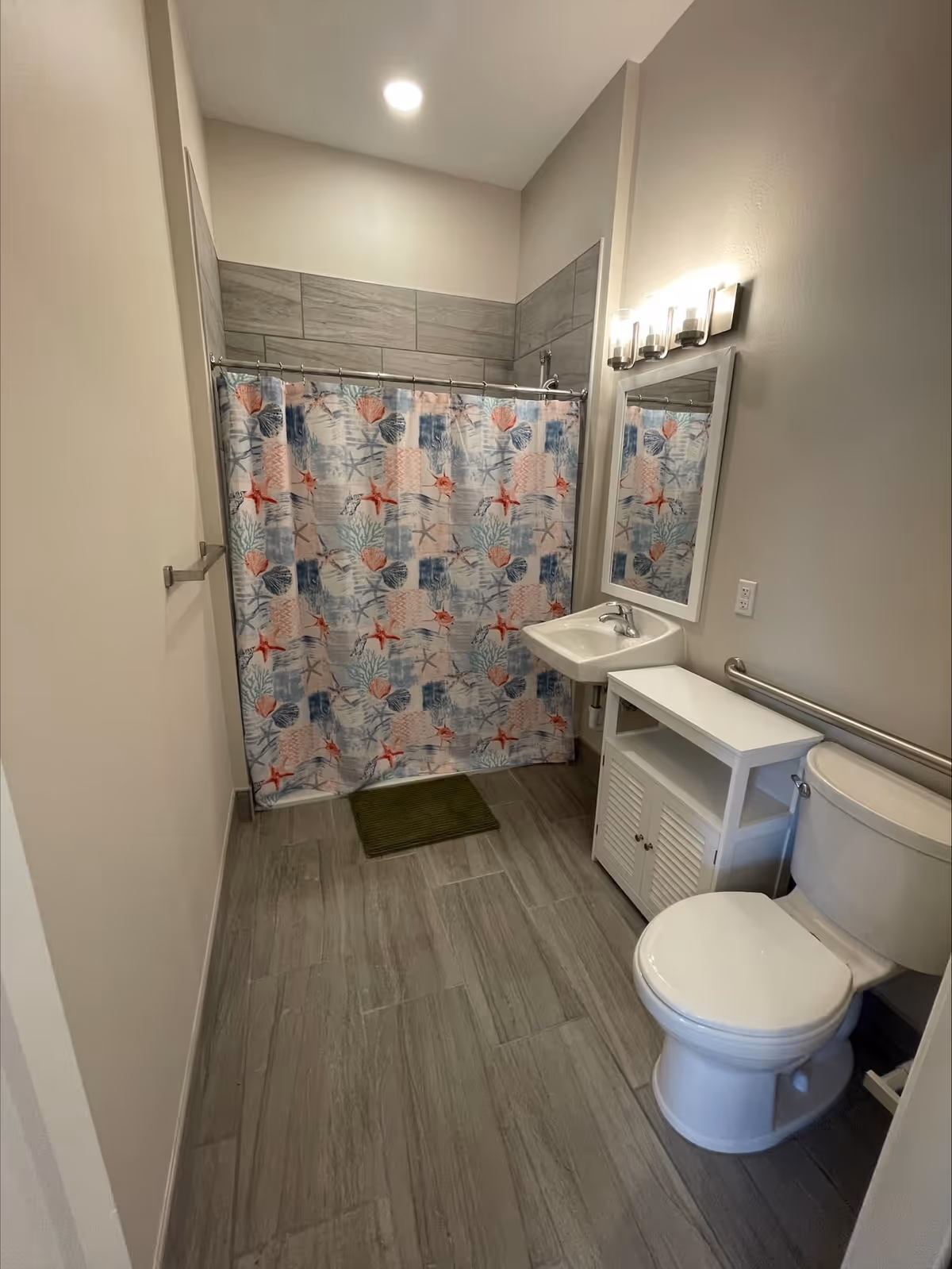 A clean bathroom with a toilet, a white sink with a mirror above it, a white cabinet next to the sink, and a shower area with a colorful shower curtain featuring seashell and starfish patterns. The floor is tiled in gray, and there is a green bath mat in front of the shower.