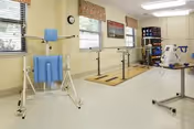 A rehabilitation room with exercise equipment including parallel bars for walking practice, a blue padded support device, and a stationary exercise bike. The room has large windows with blinds, light-colored walls, and cabinets for storage.