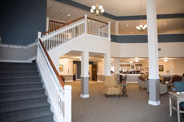 Interior view of a senior living facility showing a spacious common area with carpeted floors, a staircase with white railings and wooden handrails leading to an upper level, several armchairs and small tables arranged for seating, and multiple ceiling light fixtures providing warm lighting.