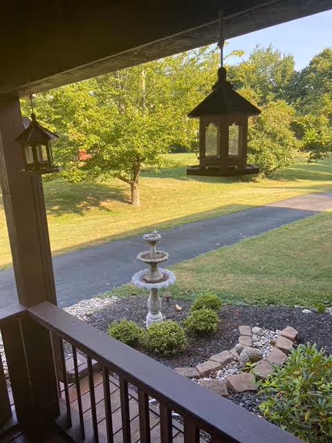 View from a covered porch overlooking a garden area with a bird feeder hanging from the porch roof, a stone birdbath surrounded by small bushes, a paved pathway, and green trees in the background under a clear sky.
