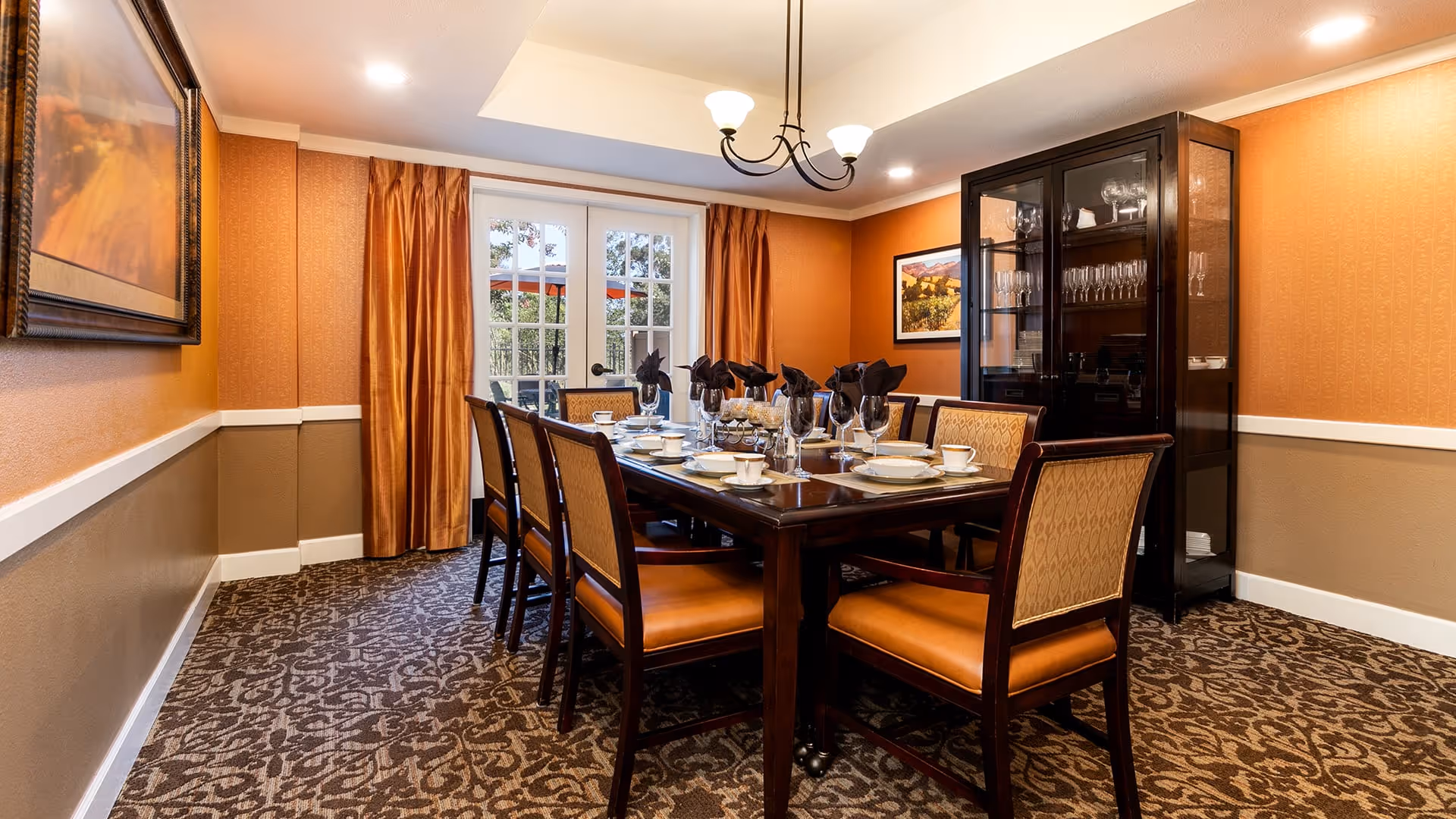 A formal dining room with a long wooden table set for a meal, surrounded by eight chairs with cushioned seats and backs. The room has patterned carpet, orange and beige walls, a glass cabinet displaying glassware, framed artwork on the walls, and a chandelier hanging from the ceiling. French doors with orange curtains lead outside.