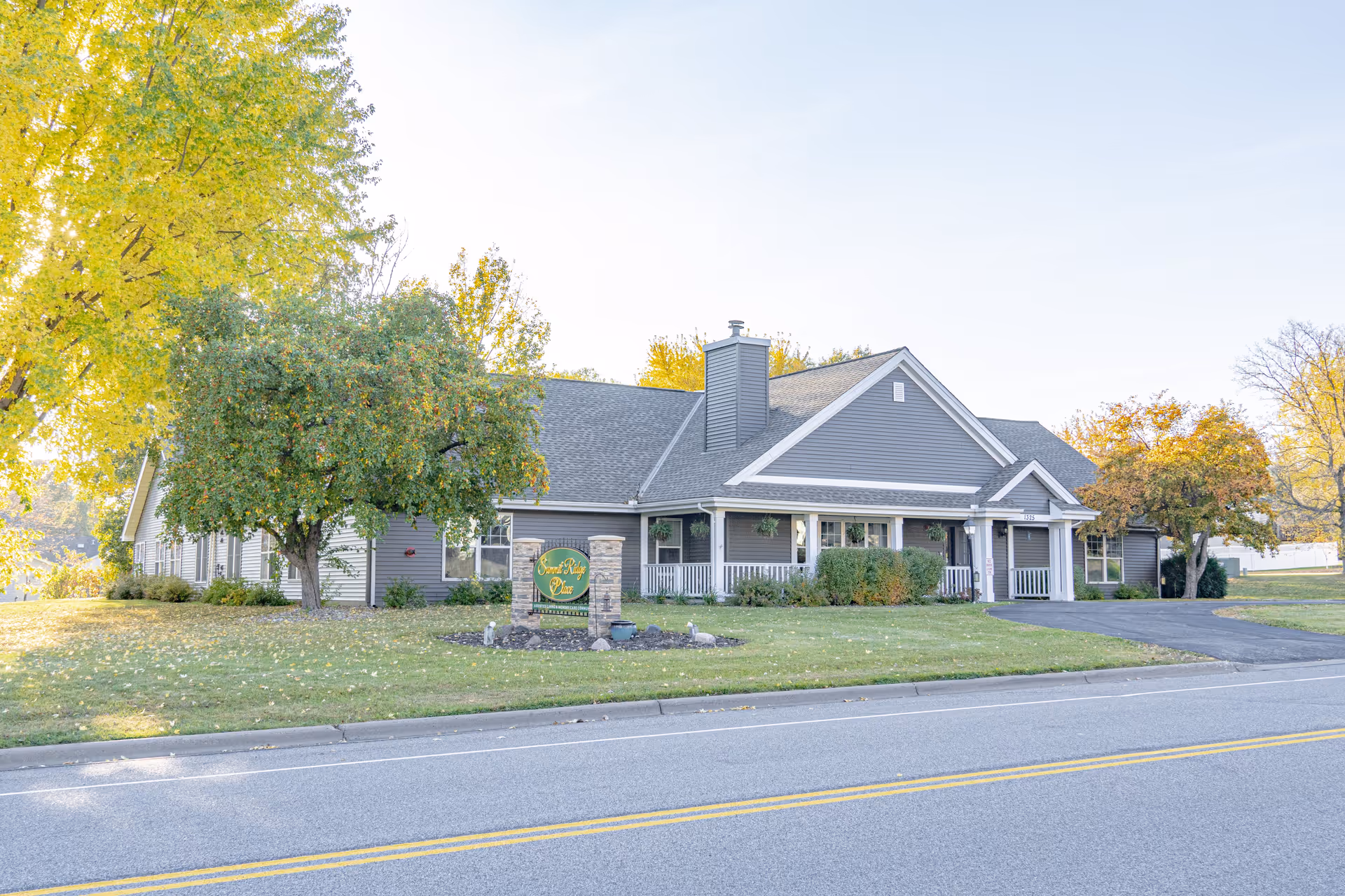 Exterior view of a single-story senior living facility building named Summit Ridge Place, surrounded by green grass and trees with autumn foliage, located next to a paved road.
