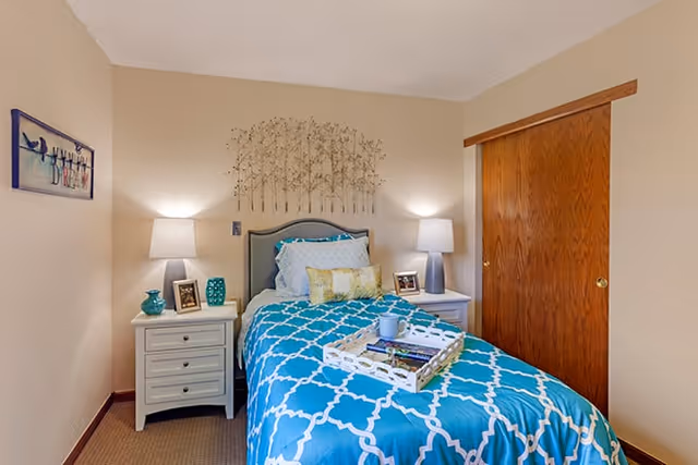 Small bedroom with a single bed dressed in blue patterned bedding, two nightstands with lamps, wall art, and a wooden closet door.