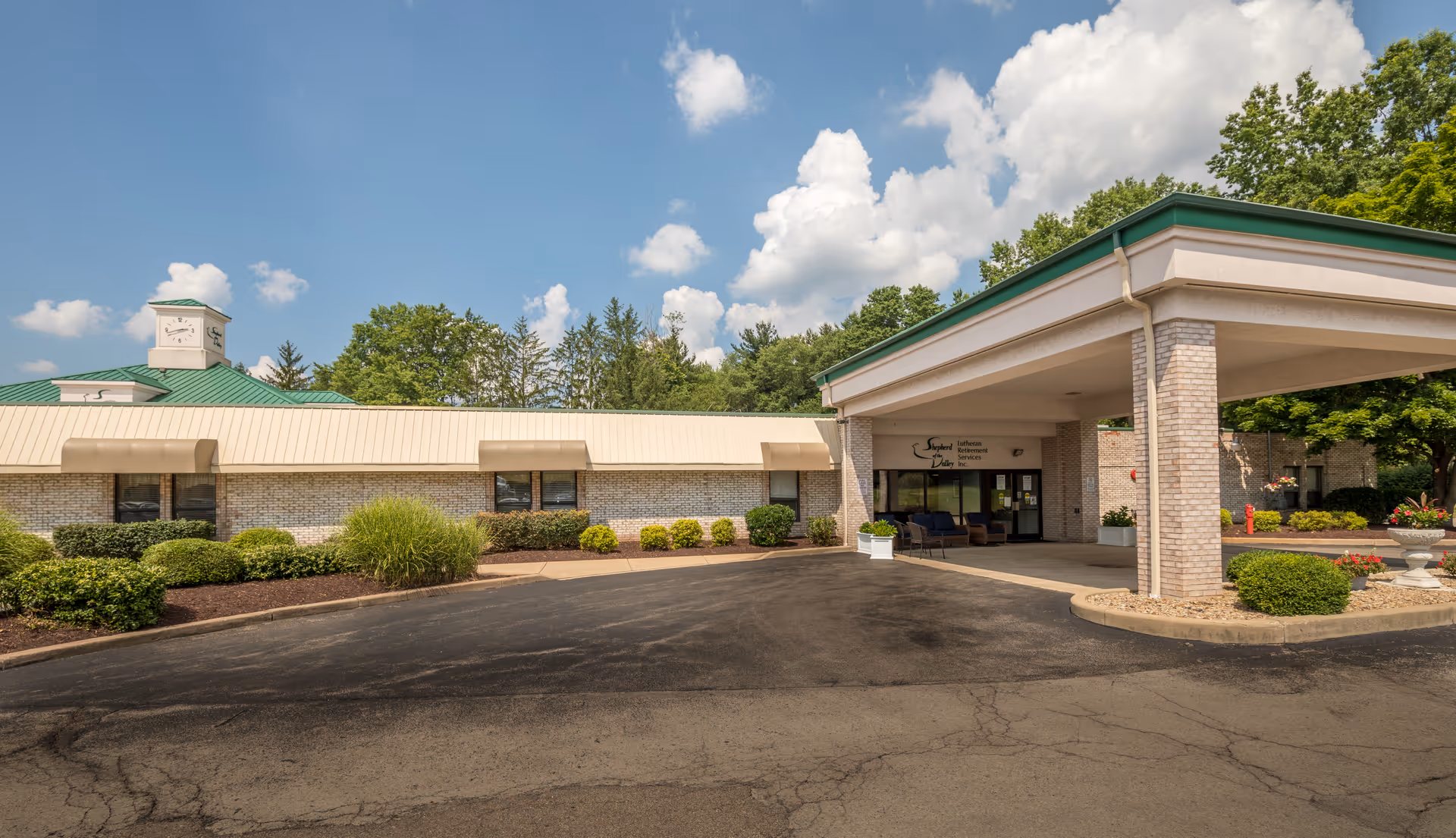 Exterior view of Shepherd of the Valley - Boardman facility showing a single-story building with a covered entrance, beige brick walls, green roof accents, and landscaped bushes and trees under a partly cloudy sky.