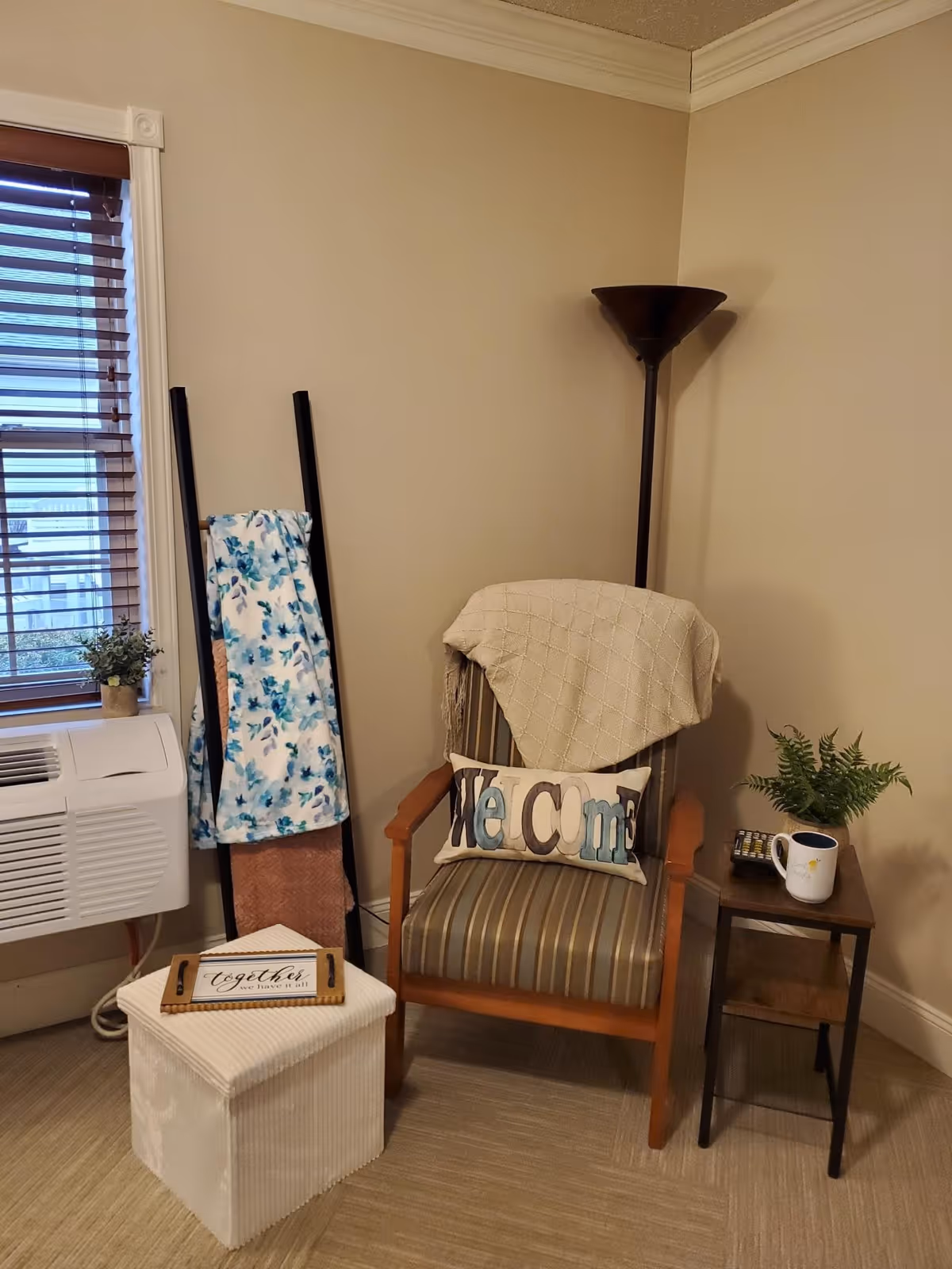A cozy corner with a striped armchair draped with a beige blanket and a decorative pillow that says 'Welcome'. Next to the chair is a small wooden side table holding a potted fern, a coffee mug, and a remote control. A white cube-shaped ottoman with a wooden tray and a sign that reads 'Together is our favorite place to be' is in front of the chair. Behind the ottoman is a ladder-style blanket rack with two blankets, one floral and one solid color. A window with wooden blinds and an air conditioning unit is on the left side, and a tall floor lamp stands in the corner.