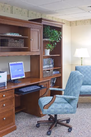 A cozy common-area office with a wooden desk and hutch holding a computer, books and a plant, a cushioned swivel chair, and a lamp-lit seating area.