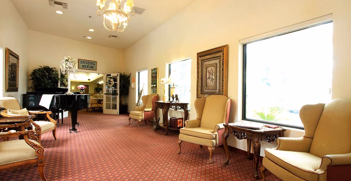 A well-lit sitting area with upholstered armchairs and wooden side tables arranged along a wall with large windows. A black grand piano is positioned near the entrance to a dining room, which is indicated by a sign above the doorway. The room has a patterned carpet and framed artwork on the walls.
