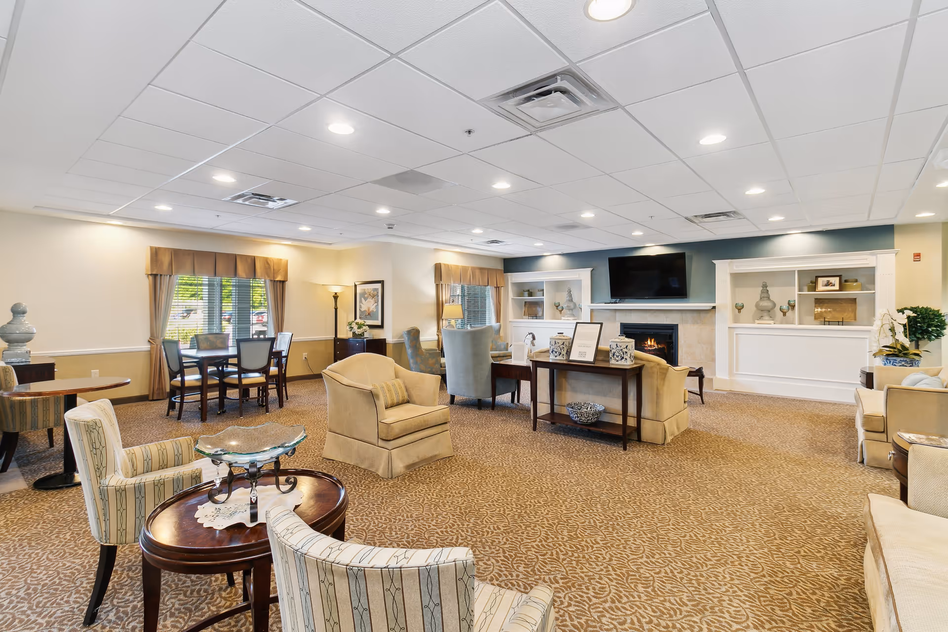 A spacious and well-lit living room area in The Landing Of Brighton facility featuring comfortable armchairs and sofas arranged around a central table. There is a fireplace with a TV mounted above it, built-in shelves with decorative items, and windows with curtains letting in natural light. The room has a patterned carpet and recessed ceiling lights.