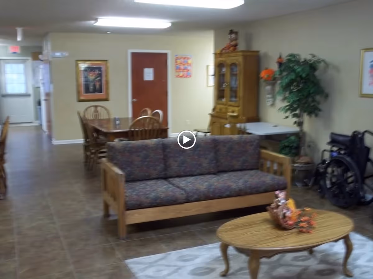Interior view of a common area in a senior living facility featuring a wooden couch with patterned cushions, a wooden coffee table with a decorative centerpiece, a dining table with chairs, a wooden cabinet, a potted plant, and a wheelchair near the wall.