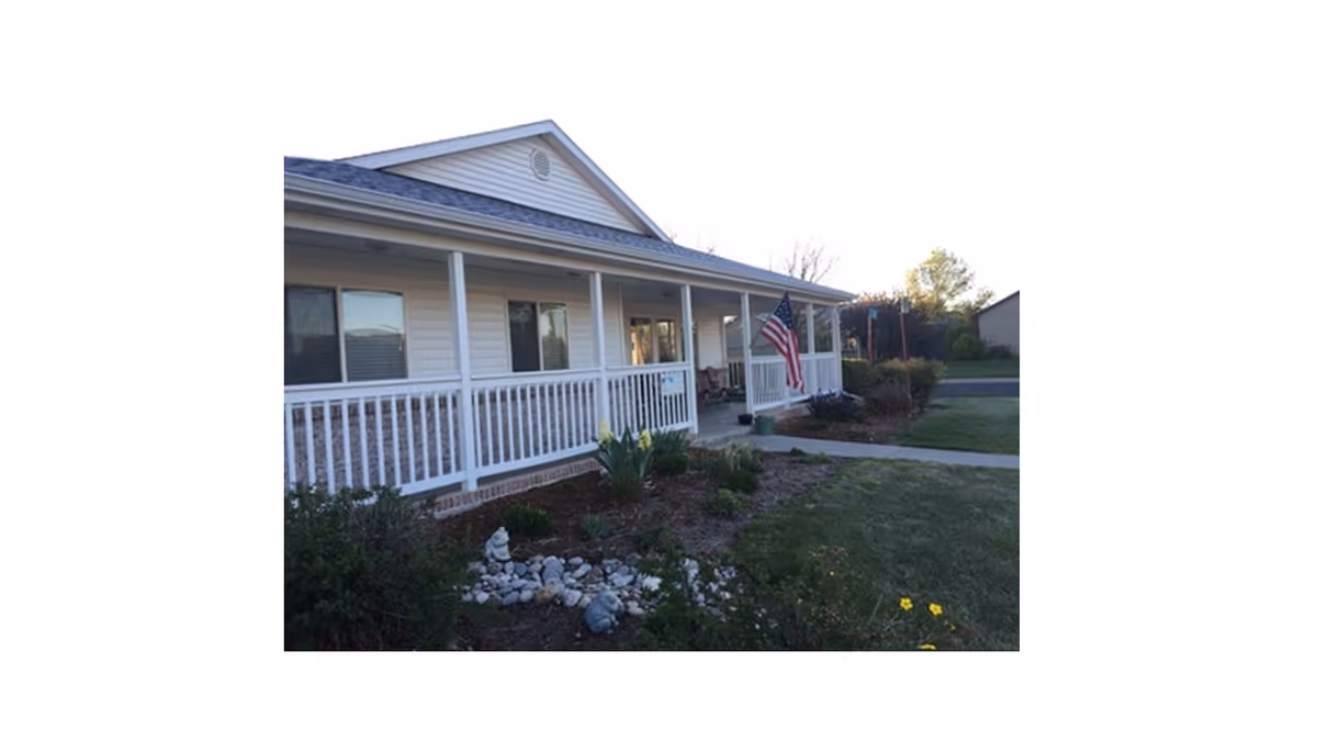 Front exterior view of a single-story building with a covered porch, white railing, and an American flag hanging near the entrance. There is a landscaped garden with rocks and plants in front of the porch.