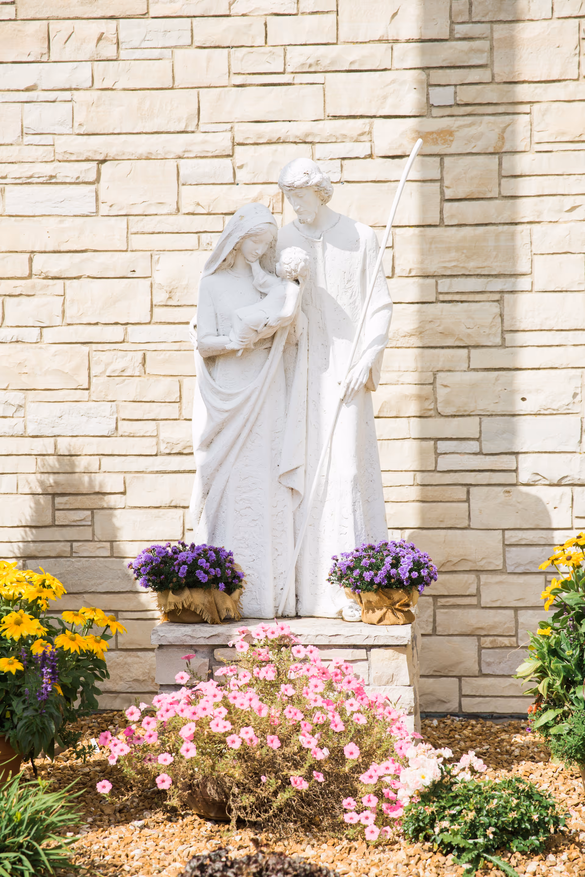 White stone statue of a family group holding a child displayed against a beige brick wall, surrounded by potted purple and pink flowers.