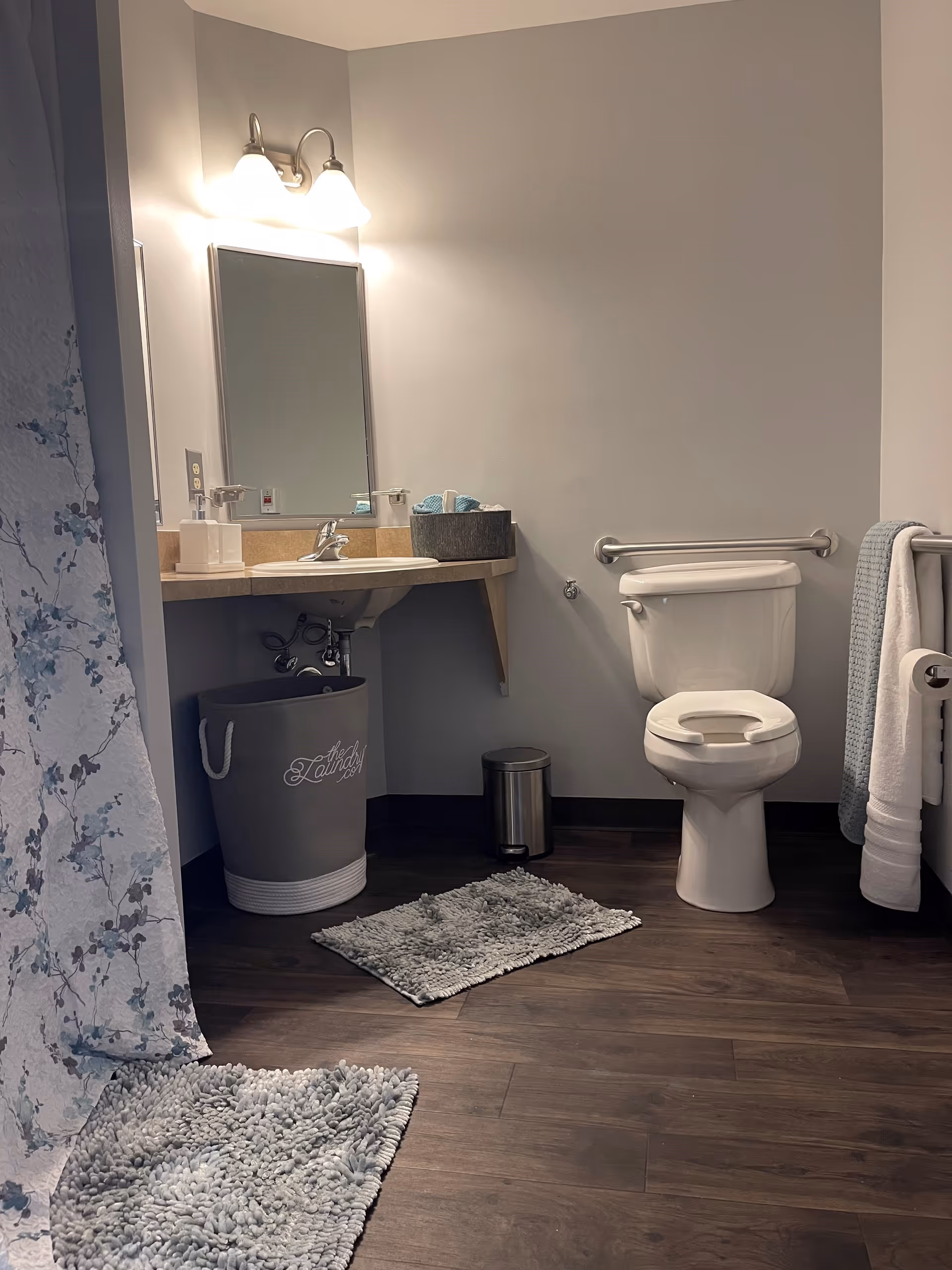 A clean and accessible bathroom with a white toilet equipped with a grab bar, a sink with a mirror and two wall-mounted lights above it, a gray laundry basket, a small trash can, two gray textured bath mats on a dark wood floor, and a floral-patterned shower curtain partially visible on the left.