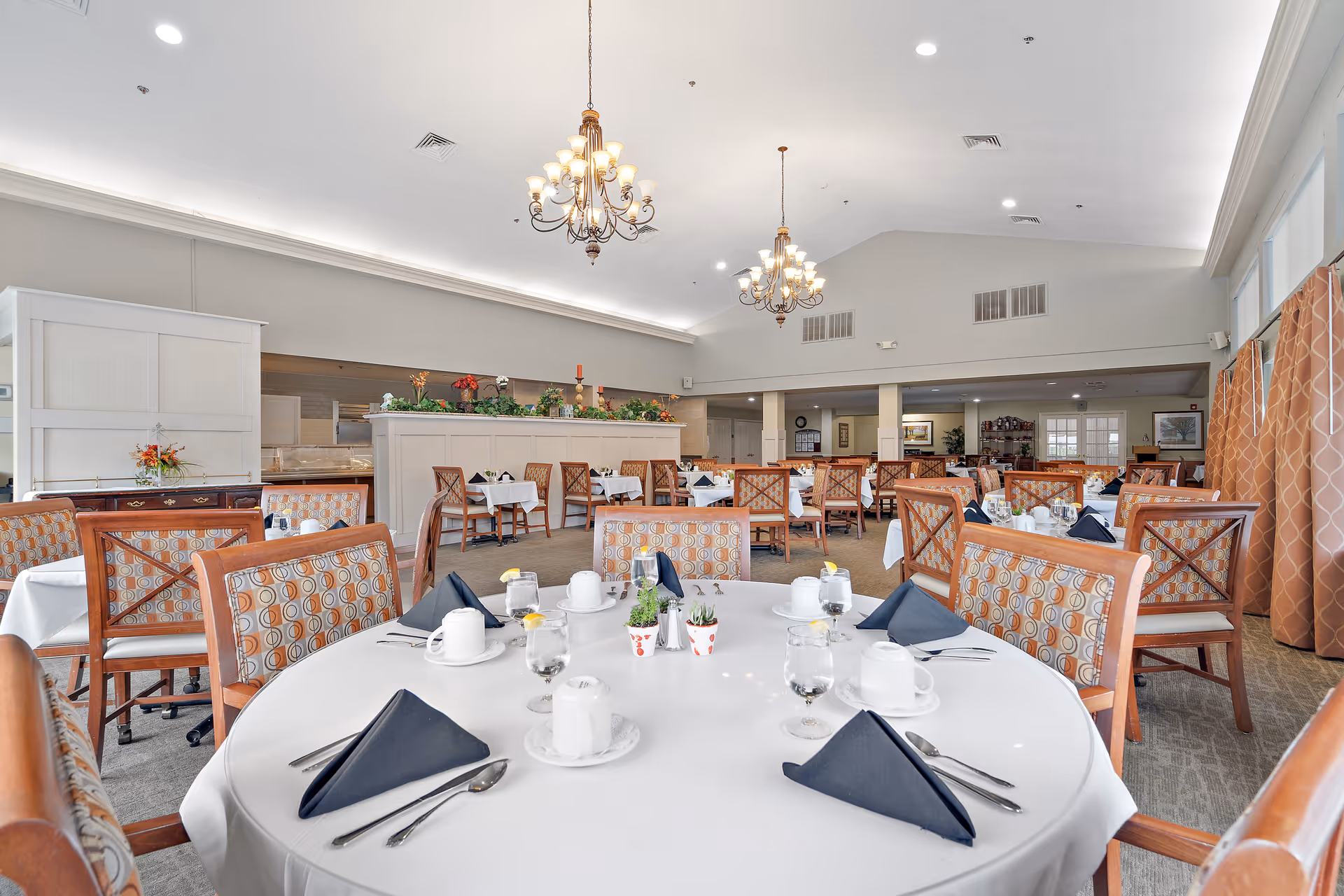 A spacious dining room with multiple round tables covered with white tablecloths, each set with black folded napkins, white cups, glasses of water with lemon, and silverware. The chairs have wooden frames with patterned upholstery. The room has high ceilings with two chandeliers, large windows with patterned curtains, and decorative plants on a partition wall.
