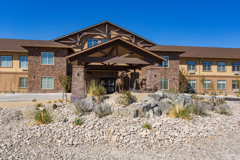 Front exterior view of Volante Senior Living of Reno building with a covered entrance, stone facade, multiple windows, and a landscaped rock garden featuring sculptures of two rams under a clear blue sky.