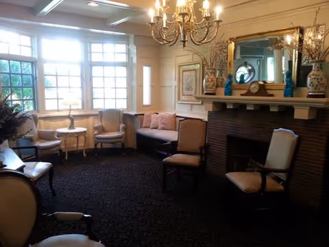 A cozy living room with several upholstered chairs and a small sofa arranged around a fireplace. The room features a large window letting in natural light, a chandelier hanging from the ceiling, and decorative items on the mantel including vases and a clock. The walls are adorned with framed artwork and a large mirror above the fireplace.