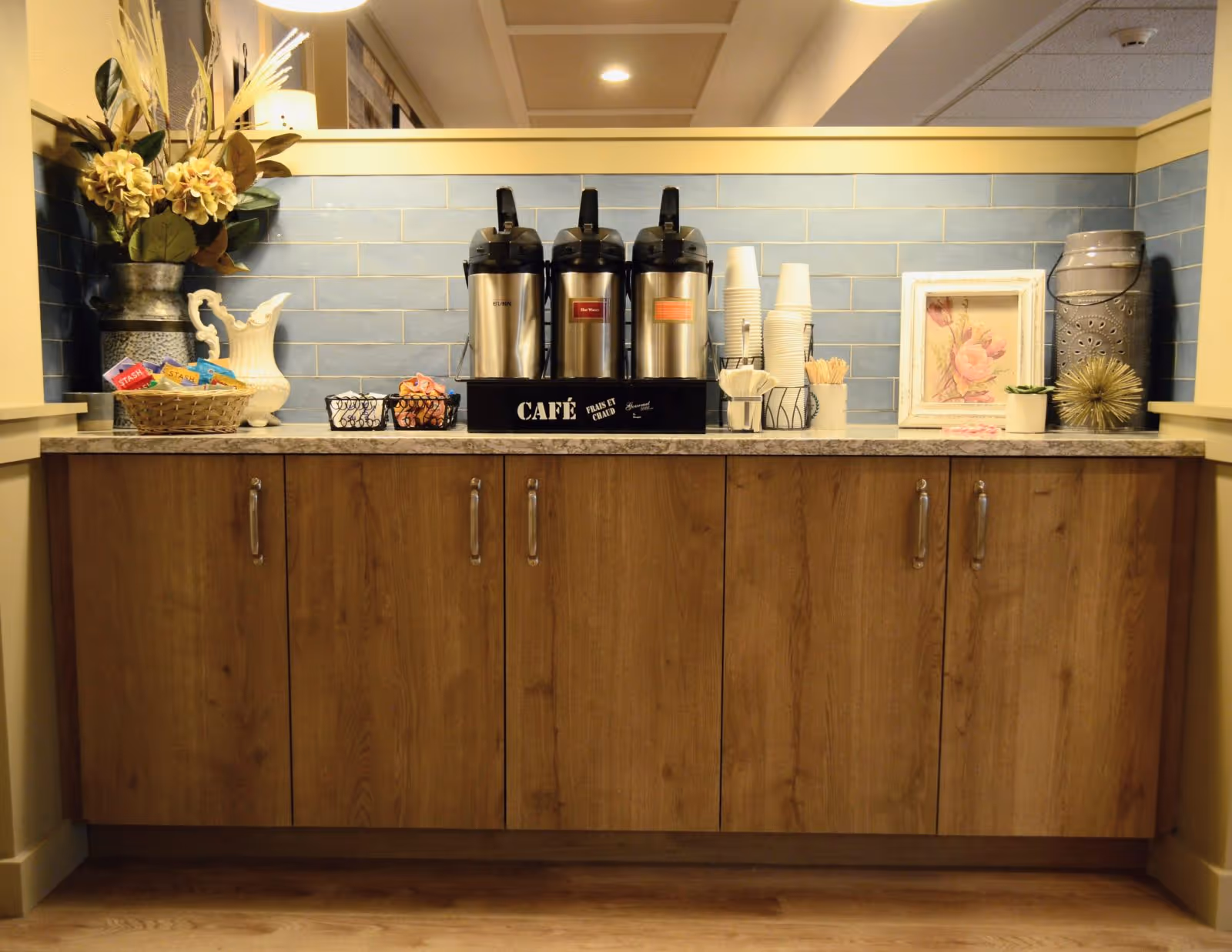 A coffee station with three stainless steel airpots labeled for different beverages, disposable cups, napkins, stirrers, and a basket of tea bags on a countertop with wooden cabinets below. The backsplash is made of light blue tiles, and decorative items including a vase with flowers, a framed floral picture, and ornamental pieces are placed on the counter.