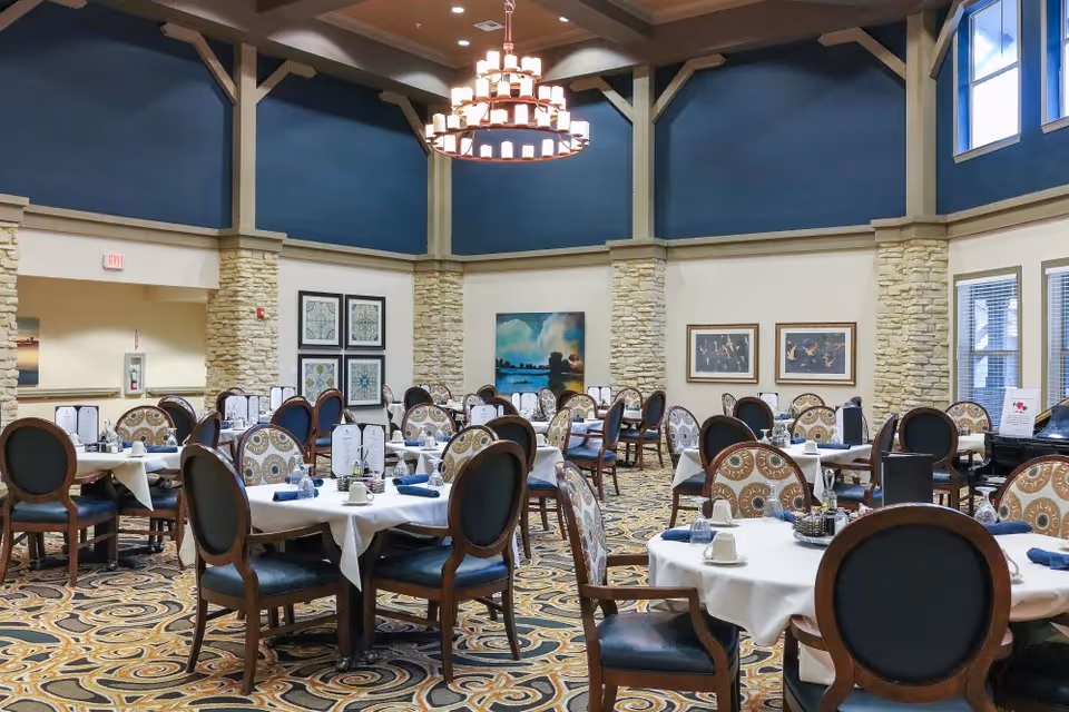 A spacious dining room with round tables covered in white tablecloths, each set with blue napkins, cups, and menus. The room features high ceilings with a large chandelier, stone pillars, patterned carpet, and framed artwork on the walls.