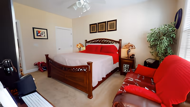 A bedroom with a wooden bed frame featuring intricate carvings, a bed covered with a red blanket and a light-colored bedspread. There is a red-covered sofa to the right, a large green plant near the window, and two bedside tables with decorative lamps. The walls are light-colored with framed artwork hanging above the bed and on the adjacent wall. A ceiling fan is visible above.