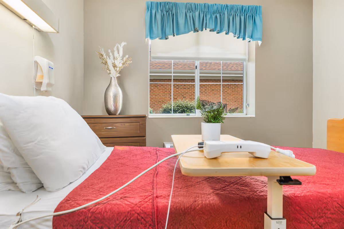 A hospital-style bed with white pillows and a red blanket in a room. A wooden bedside table with a silver vase holding dried flowers is next to the bed. A window with a blue valance shows a brick building outside. A small table on wheels with a white telephone and a potted plant is positioned over the bed.