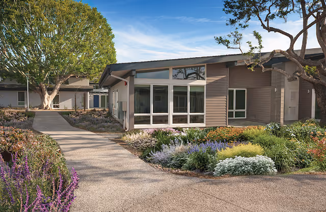 Exterior view of a single-story building at Wesley Palms Retirement Community with large windows, surrounded by landscaped gardens featuring various shrubs and flowering plants, and a paved walkway leading to the entrance under a clear blue sky.