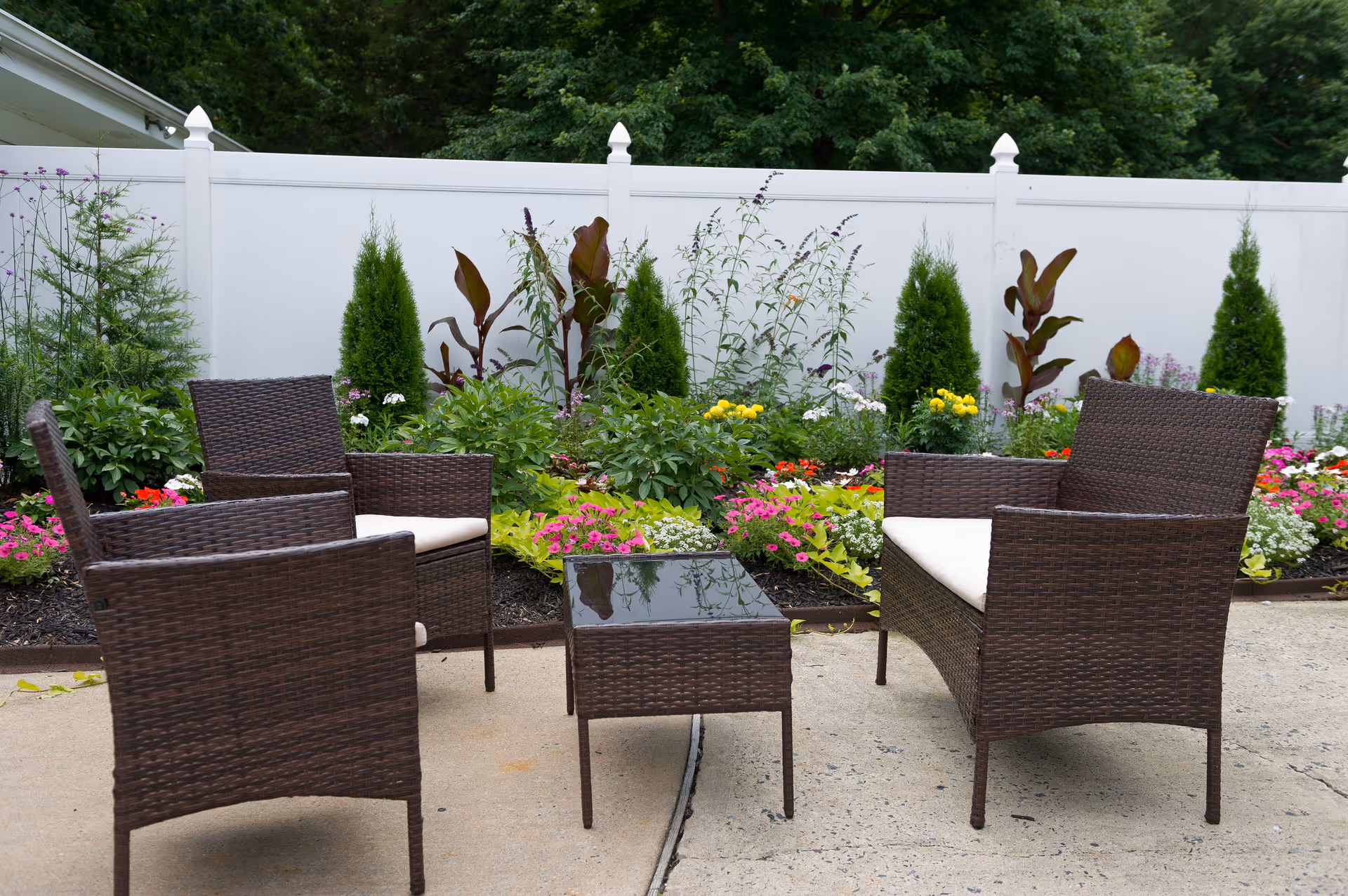 Outdoor patio area with four brown wicker chairs with white cushions arranged around a small rectangular glass-top table. Behind the seating area is a garden bed filled with colorful flowers and green shrubs, bordered by a white fence and dense trees in the background.