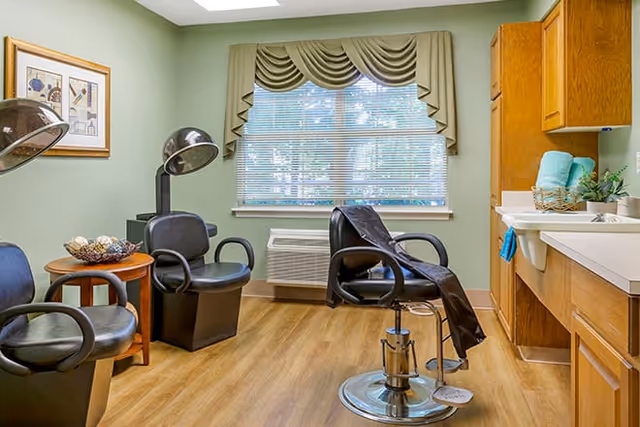 Salon-style room with black styling chairs, hooded hair dryers, a barber chair, sink and wooden cabinets beneath a window with a valance.
