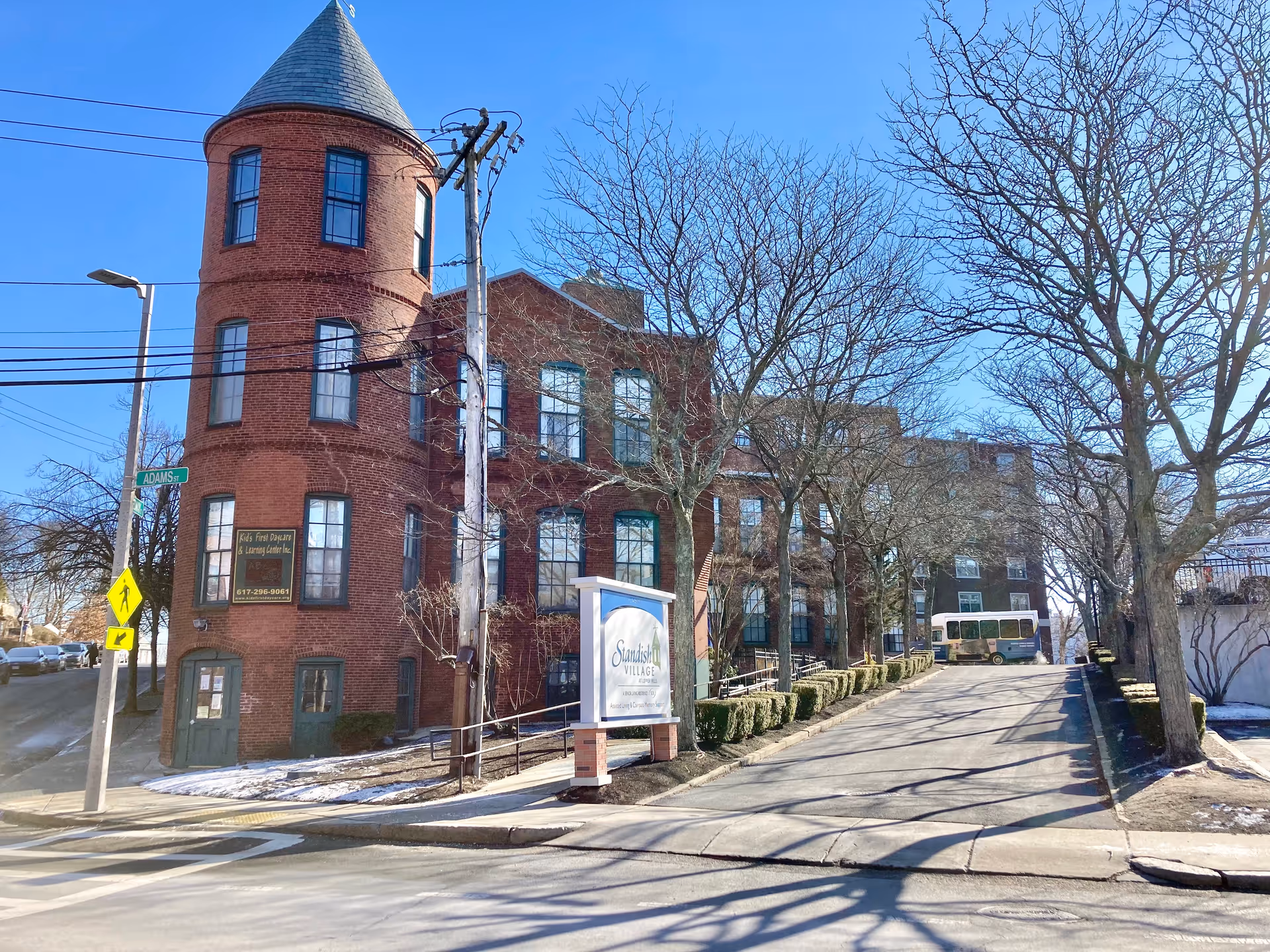 Red brick multi-story building with a rounded tower, driveway and a 'Standish Village' sign at the entrance.