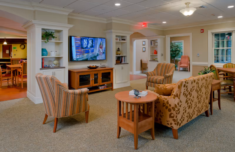 A cozy senior living common area with patterned armchairs and a sofa arranged around a wooden coffee table. A flat-screen TV is mounted on the wall above a wooden cabinet. Built-in shelves with decorative items flank the TV. The room has beige walls, carpeted floors, and recessed lighting. Adjacent rooms with additional seating and tables are visible through open doorways.