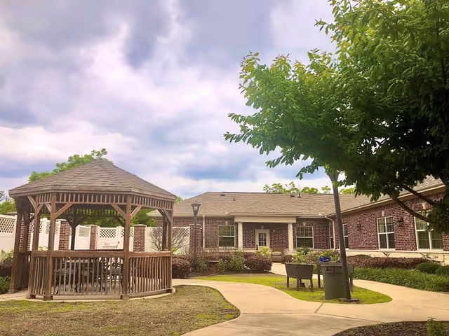 Outdoor area of a senior living facility featuring a wooden gazebo with seating, a paved walkway, green trees, and a brick building in the background under a cloudy sky.