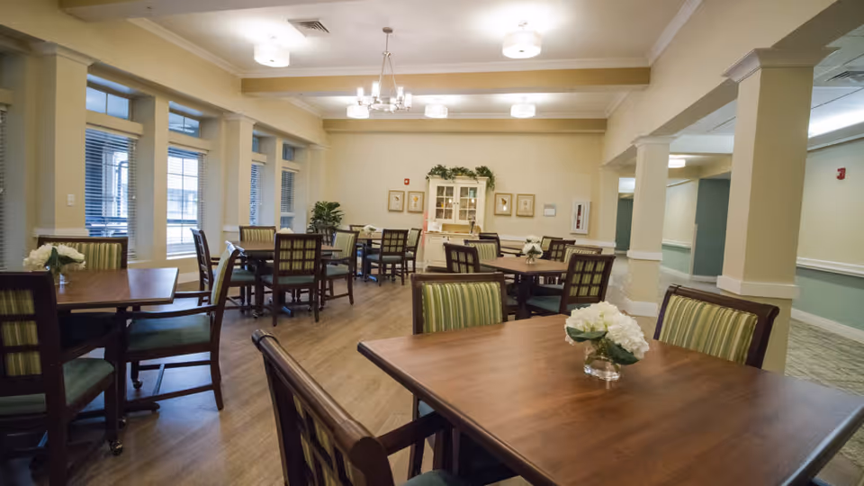 A well-lit dining room with several wooden tables and chairs arranged neatly. Each table has a small vase with white flowers. The room features large windows with blinds, light-colored walls, and ceiling lights. There is a cabinet with decorative items and framed pictures on the far wall.