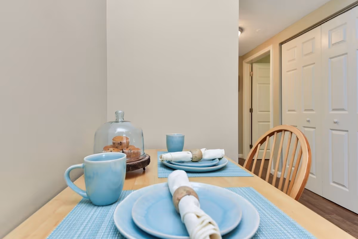 A small dining table set for two with light blue plates, matching mugs, and white napkins with napkin rings. There is a glass dome covering muffins on a wooden stand. The table has blue placemats and is positioned near a hallway with white doors and a wooden chair.