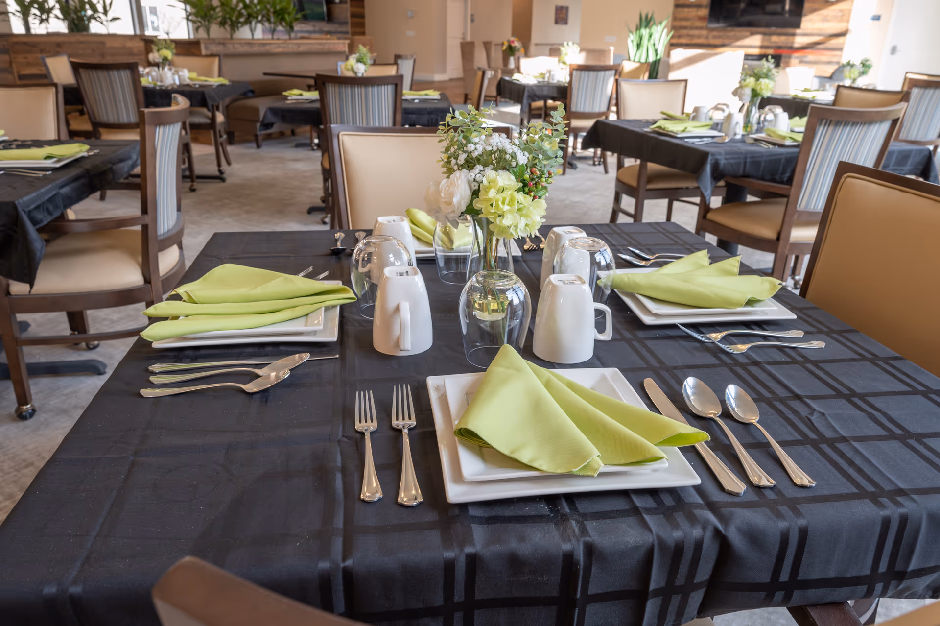 A dining room with tables set for a meal, featuring black tablecloths, neatly folded green napkins on white square plates, silverware, white mugs, and small floral centerpieces. The room has multiple tables and chairs arranged for dining, with natural light coming in from the windows.