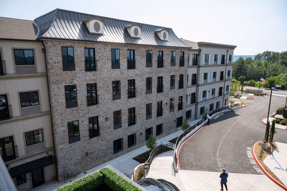 Exterior front of a multi-story stone and stucco senior living building with rows of windows and a curved driveway entrance.