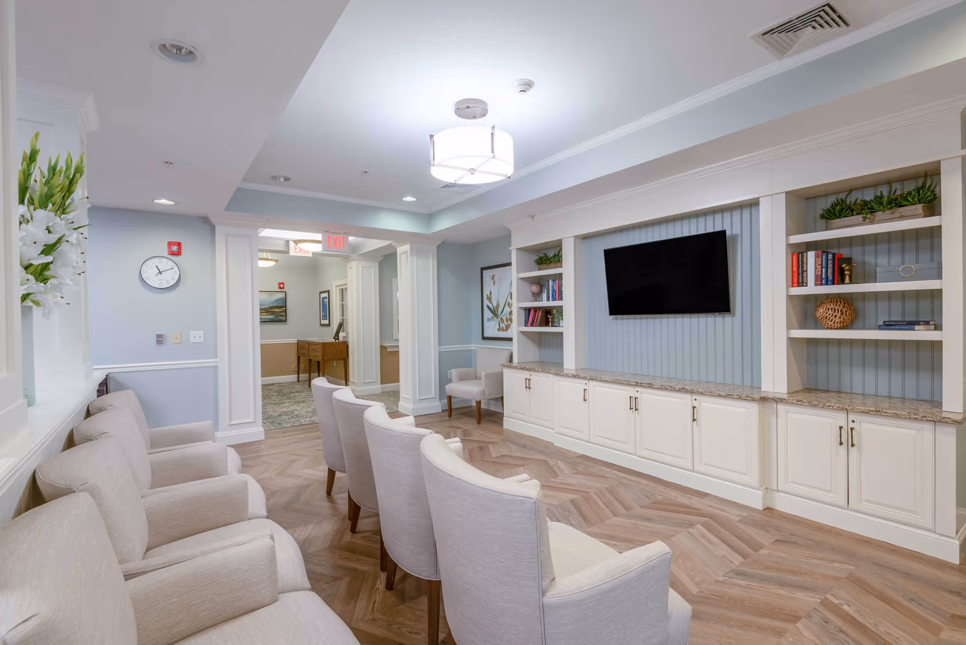 A bright and clean senior living facility common area with beige upholstered chairs arranged in rows facing a wall-mounted flat screen TV. The wall behind the TV has built-in white cabinetry and shelves decorated with books and plants. The room has light blue walls, wood-patterned flooring, and a ceiling light fixture. A clock and floral arrangement are visible on the left wall, and an exit sign is seen in the background hallway.