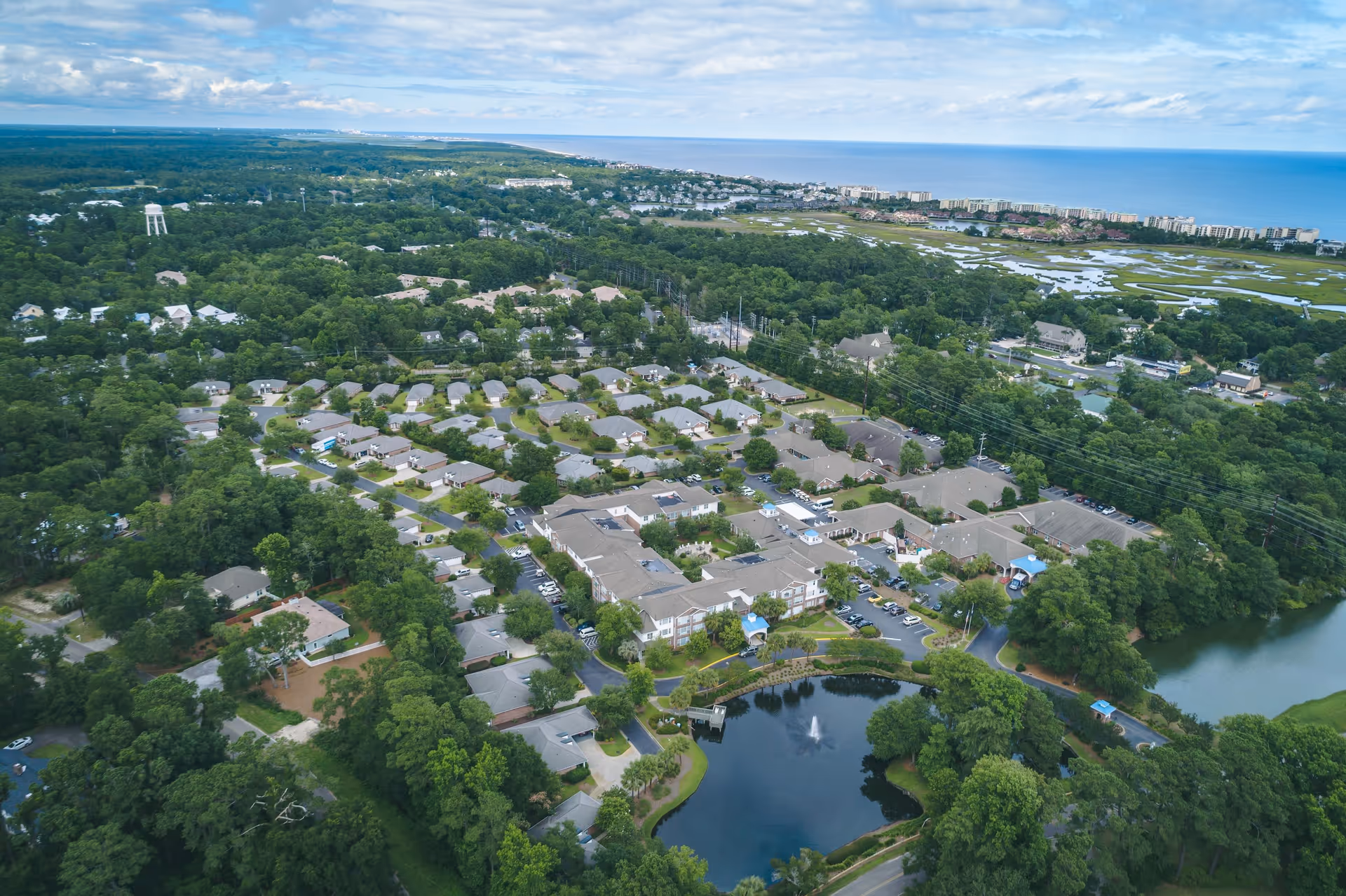 Aerial view of The Lakes at Litchfield senior living facility surrounded by dense green trees, with multiple buildings, parking lots, a pond with a fountain, and a distant view of the coastline and ocean under a partly cloudy sky.