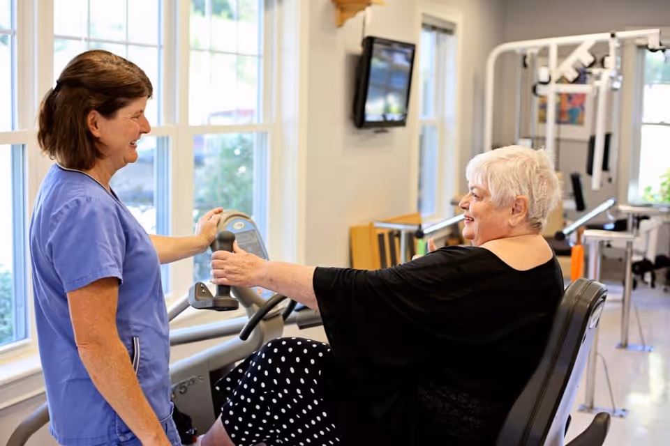 An elderly woman using a seated exercise machine in a fitness room, assisted by a healthcare worker in blue scrubs. The room has large windows letting in natural light and various exercise equipment in the background.