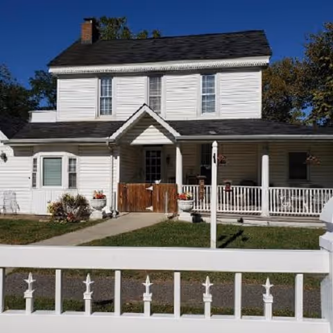 Two-story white house with a dark shingled roof, a front porch with white railings, and a small garden area. The house is viewed from behind a white fence with decorative posts, under a clear blue sky.
