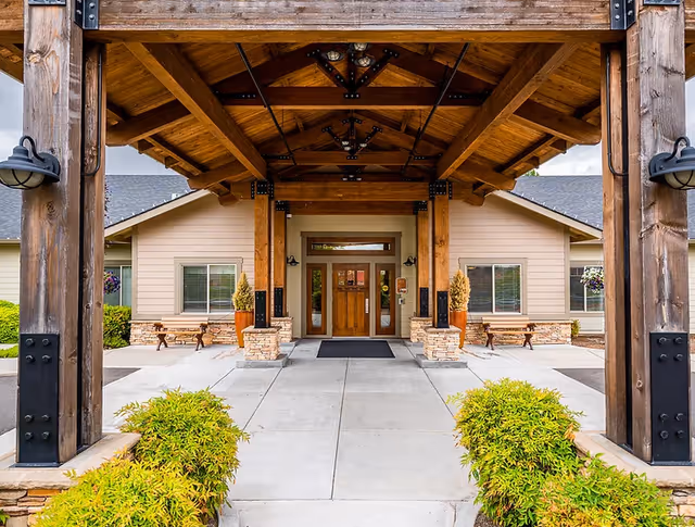 Entrance of Sunnyside Meadows Memory Care facility featuring a covered wooden pergola with exposed beams, a concrete walkway flanked by green shrubs, beige building exterior with windows, benches, and potted plants near the doorway.