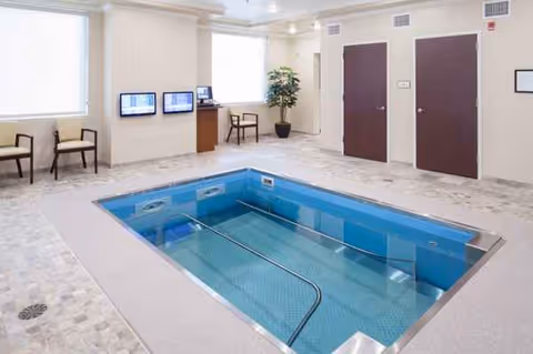 Indoor pool area of a senior living facility with seating, TVs, and a potted plant.