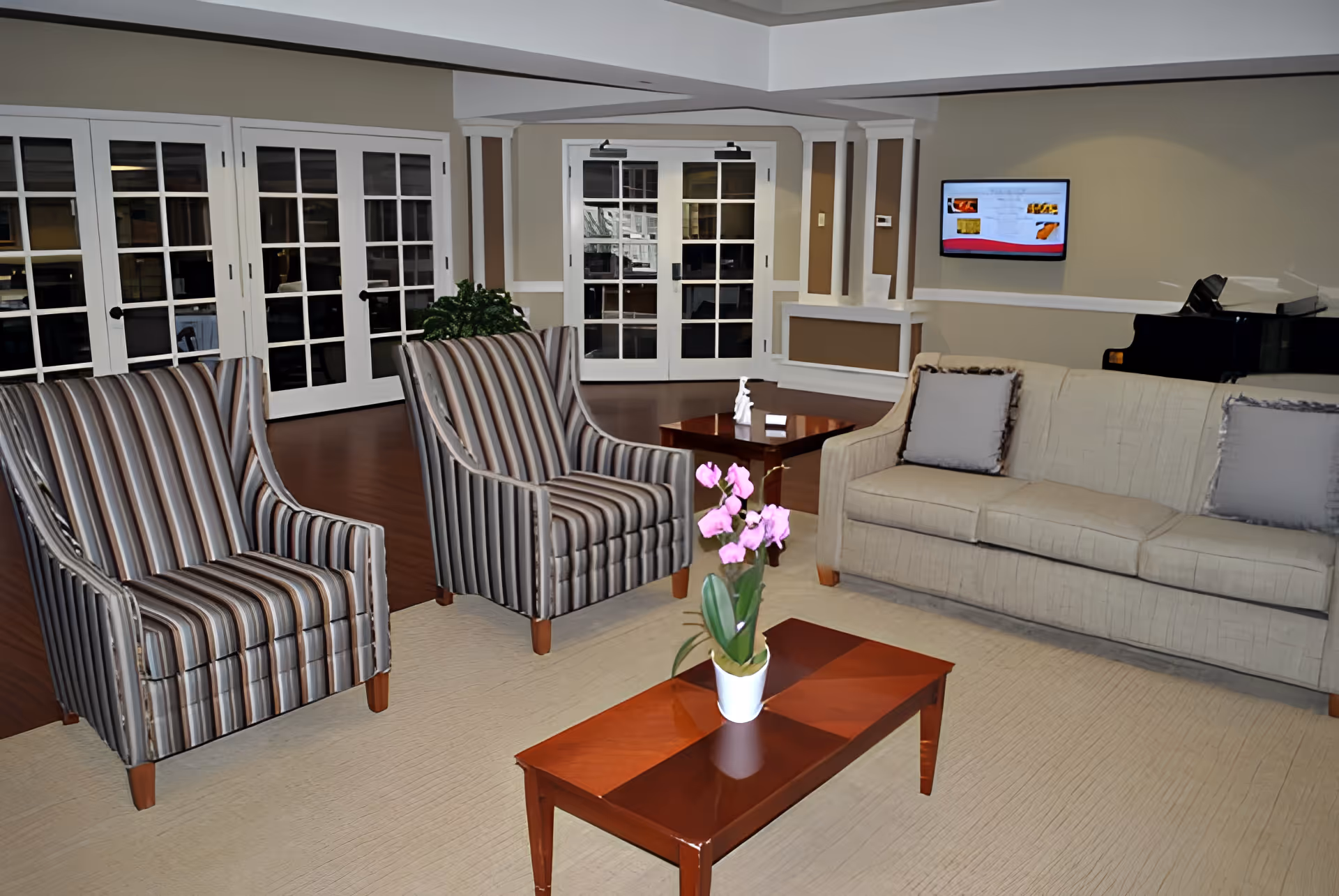Communal living room with striped armchairs, a beige sofa, coffee tables and a potted orchid in front of glass-paneled doors.