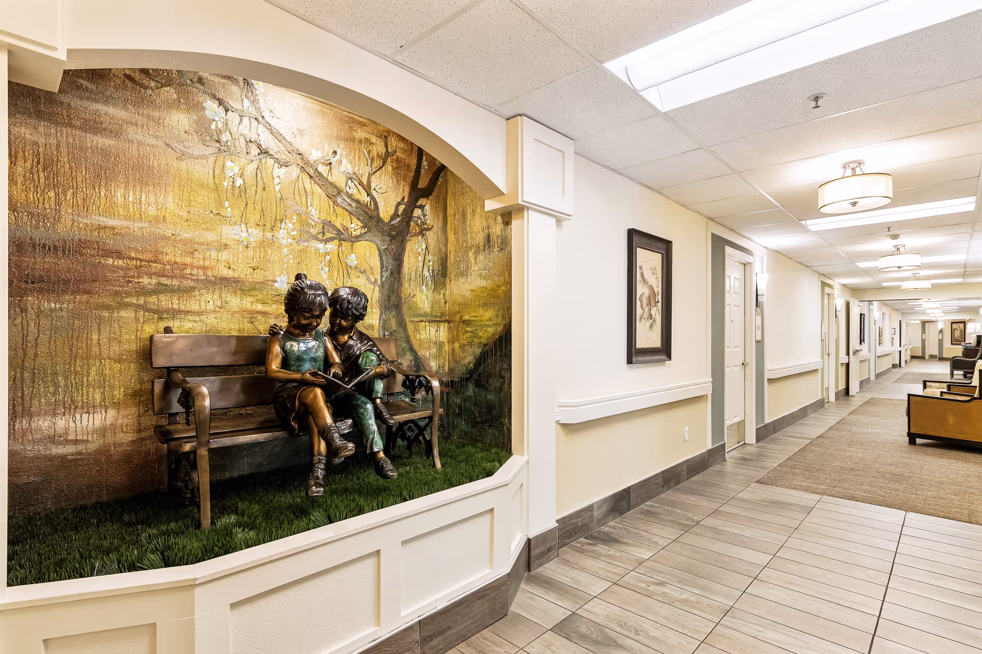 A well-lit hallway in a senior living facility with tiled floors and beige walls. On the left side, there is a decorative alcove featuring a bronze statue of two children sitting on a bench reading a book, set against a mural of a tree with blossoms. The hallway extends into the distance with doors and framed artwork along the walls, and seating areas visible further down.