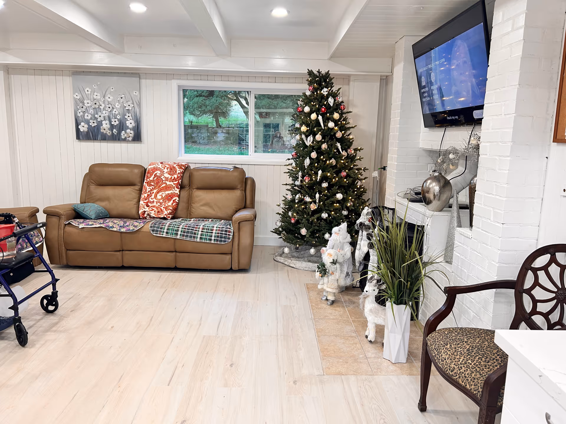 A cozy living room decorated for Christmas with a brown leather sofa adorned with colorful blankets, a decorated Christmas tree, holiday figurines including Santa Claus and reindeer, a flat-screen TV mounted on a white brick wall above a fireplace, a wooden chair with a leopard print cushion, and a walker visible on the left side.