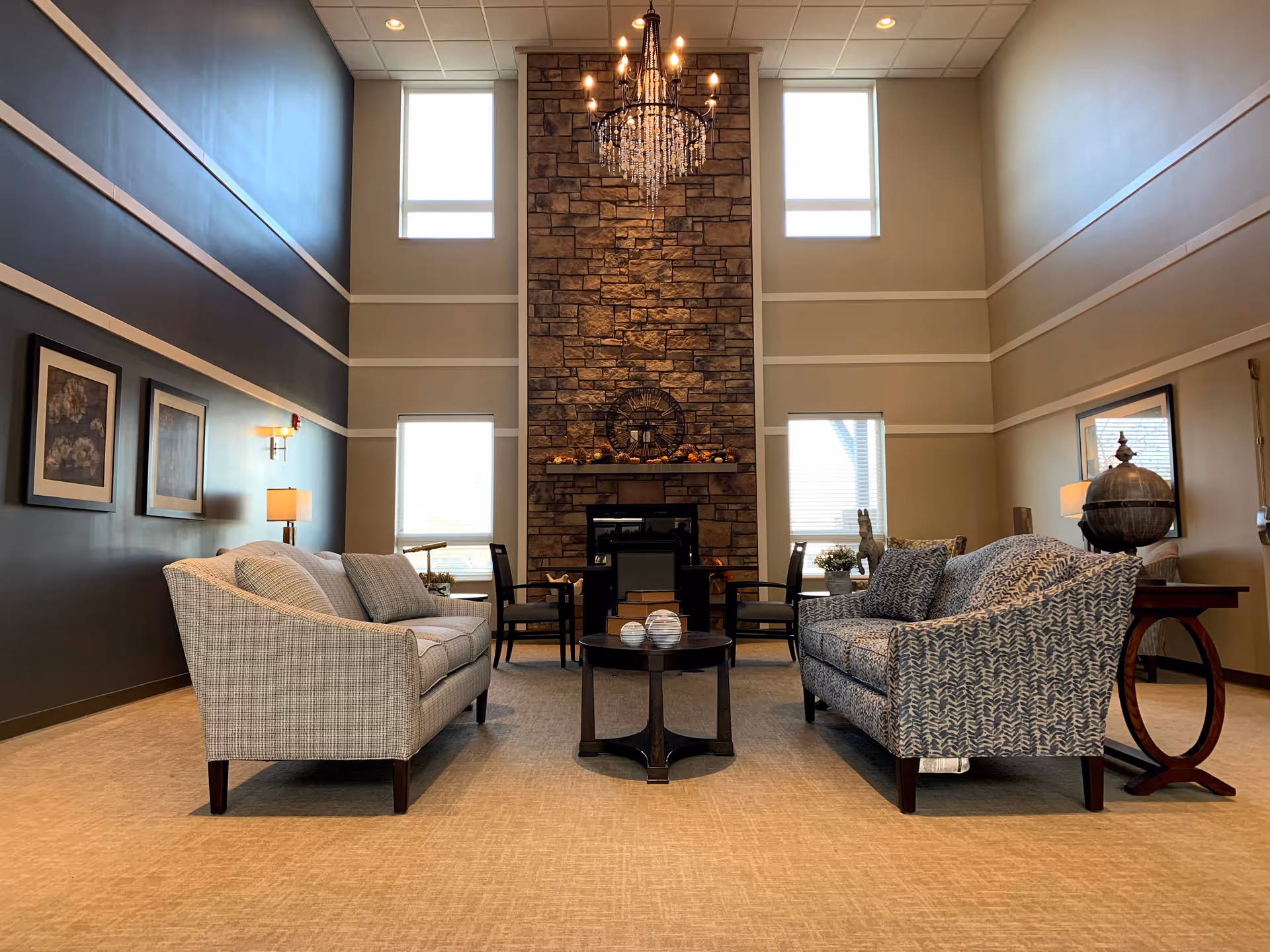 A spacious and elegant living room with two patterned sofas facing each other, a round coffee table in the center, and a tall stone fireplace with a chandelier hanging above. The room has high ceilings with large windows on either side of the fireplace, framed artwork on the walls, and side tables with lamps and decorative items.