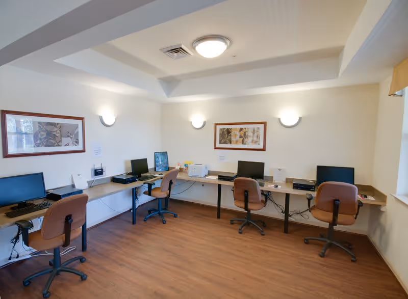 A small computer room with four workstations along two walls, each equipped with a computer monitor, keyboard, and office chair. The room has wooden flooring, white walls, two framed abstract artworks, and ceiling and wall-mounted lights providing illumination.