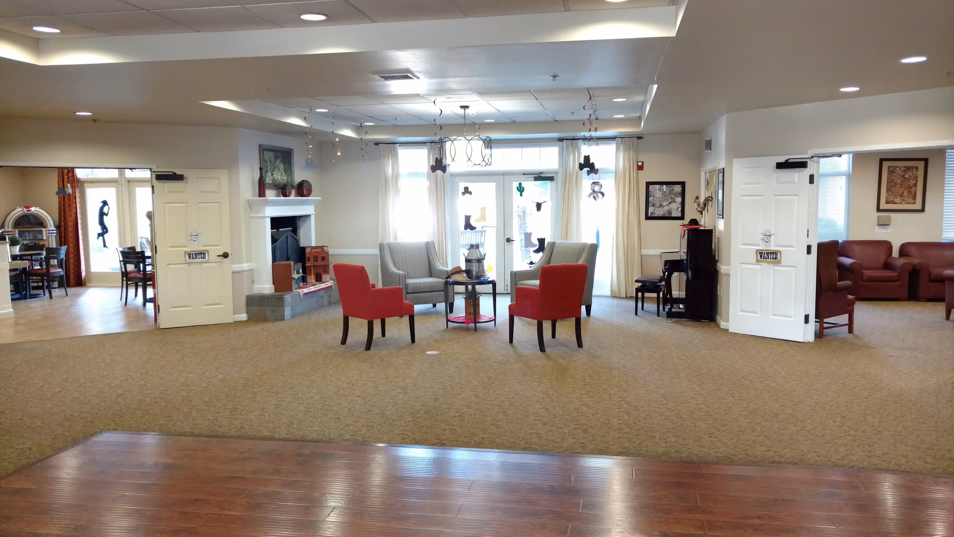 A spacious common area in a senior living facility featuring a seating arrangement with two red chairs and two gray armchairs around a small round table. The room has beige carpeting, a white fireplace with decorative items on the mantel, and large windows with sheer curtains letting in natural light. To the left, there is an open doorway leading to a dining area with tables and chairs, and to the right, another open doorway leads to a lounge area with red sofas. The ceiling has recessed lighting and decorative hanging ornaments near the windows.