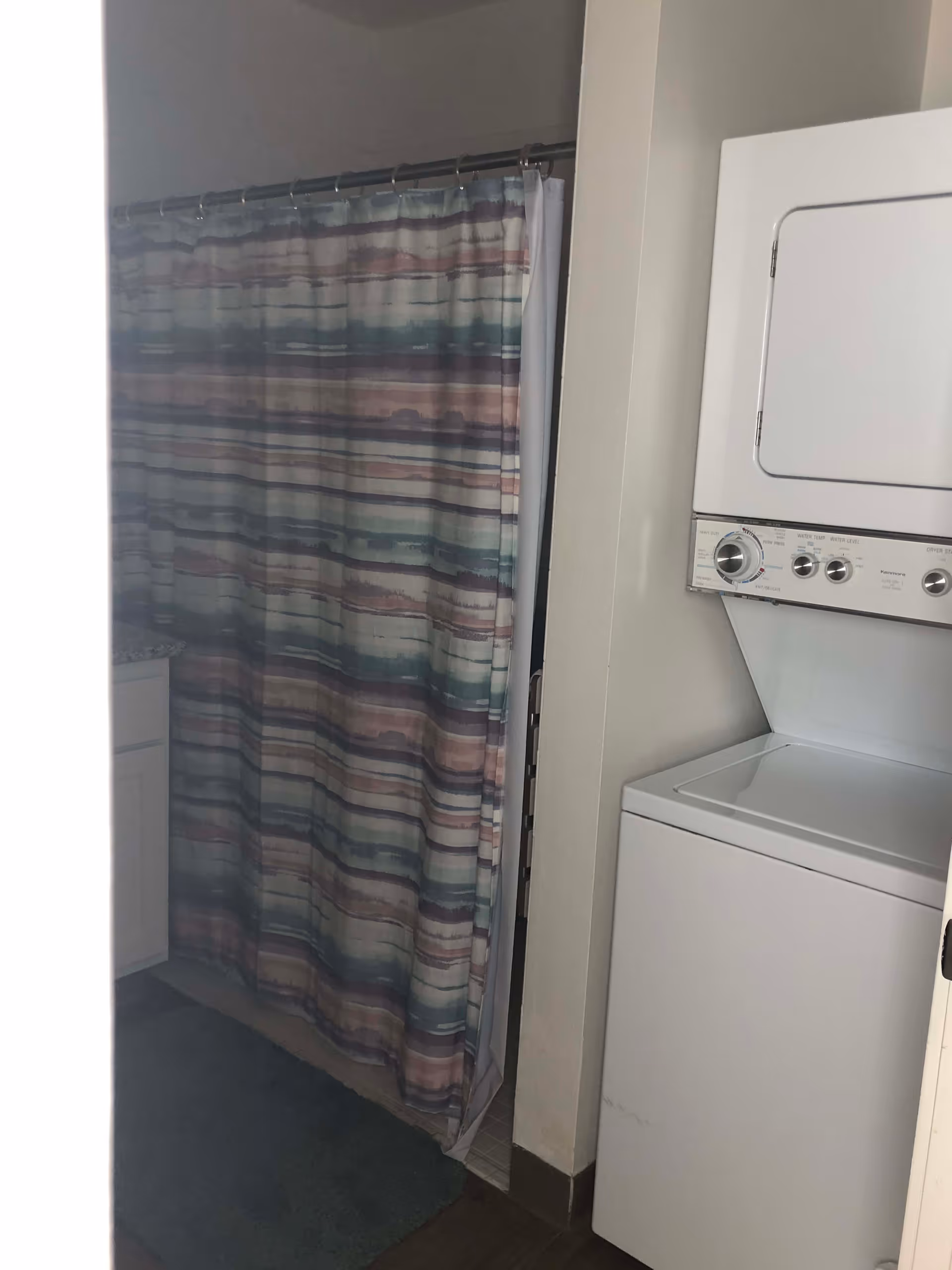 A bathroom area with a colorful striped shower curtain and a stacked white washer and dryer unit next to it. Part of a white cabinet and a blue bath mat are also visible.