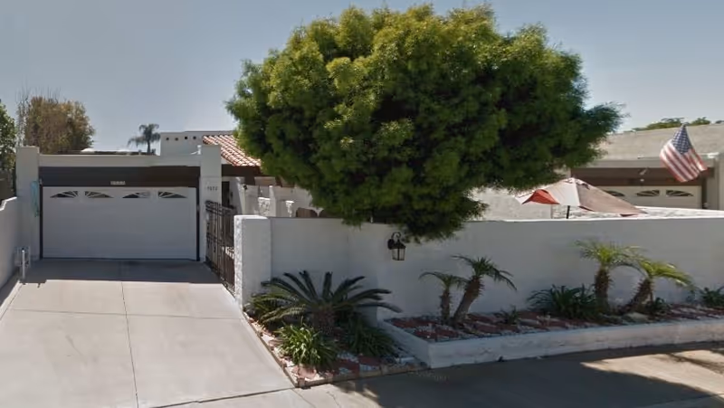 Exterior view of a single-story residential building with a white garage door, a driveway, a white wall with a mounted light fixture, a large leafy tree, and some small palm plants in a landscaped area. An American flag is visible on the right side of the image.