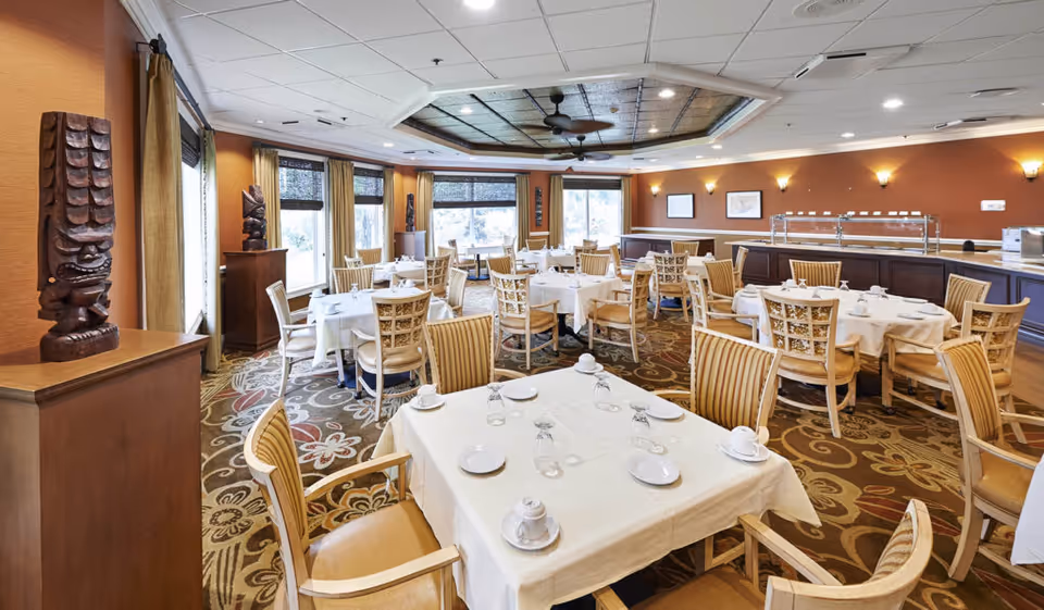 A spacious dining room with multiple round tables covered with white tablecloths, each set with cups, saucers, plates, and glasses. The room features large windows with curtains, decorative wooden sculptures on pedestals, patterned carpet, and warm-toned walls with wall sconces. A buffet counter is visible along one wall.