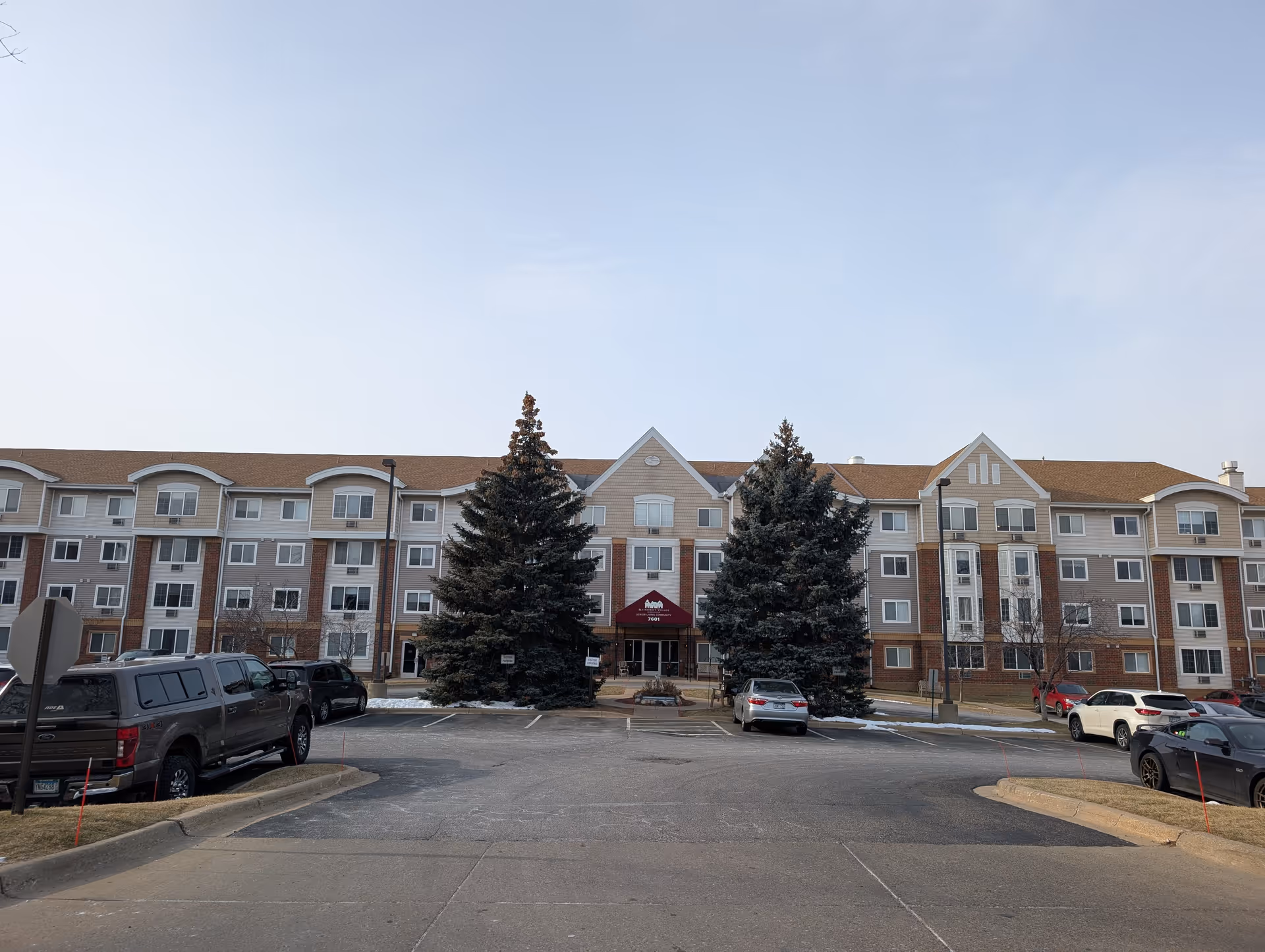 Front exterior of a multi-story senior living building with a canopy entrance, two large evergreen trees, and parked cars in the driveway.